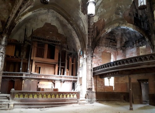 Interior of an abandoned church with crumbling walls, a wooden altar, and gothic arches, illuminated by natural light.