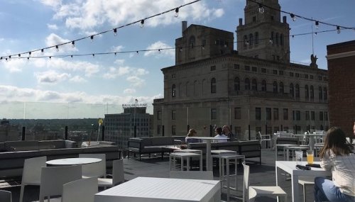 Rooftop terrace with white furniture, string lights, and a historic building in the background under a partly cloudy sky.