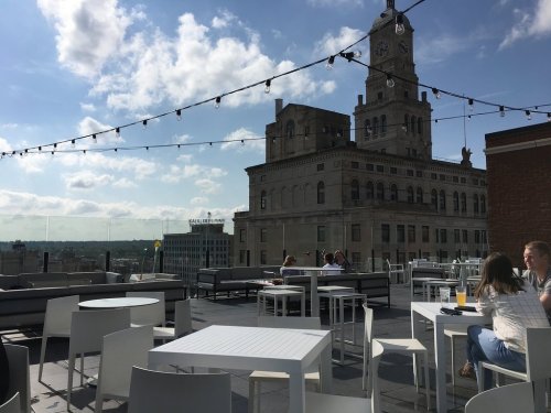 Rooftop terrace with white furniture, string lights, and a view of a historic building under a partly cloudy sky.