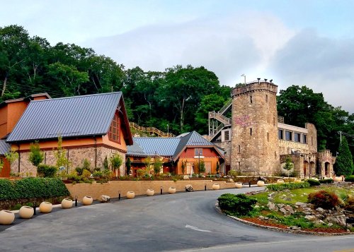 A stone castle-like building next to a modern structure, surrounded by greenery and a winding road.