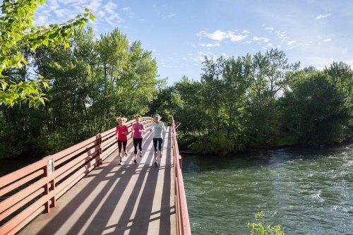 Three people walk joyfully on a bridge over a river, surrounded by lush greenery and a clear blue sky.
