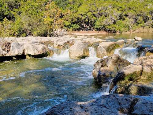 A serene river flows over smooth rocks, surrounded by lush green trees and a clear blue sky.