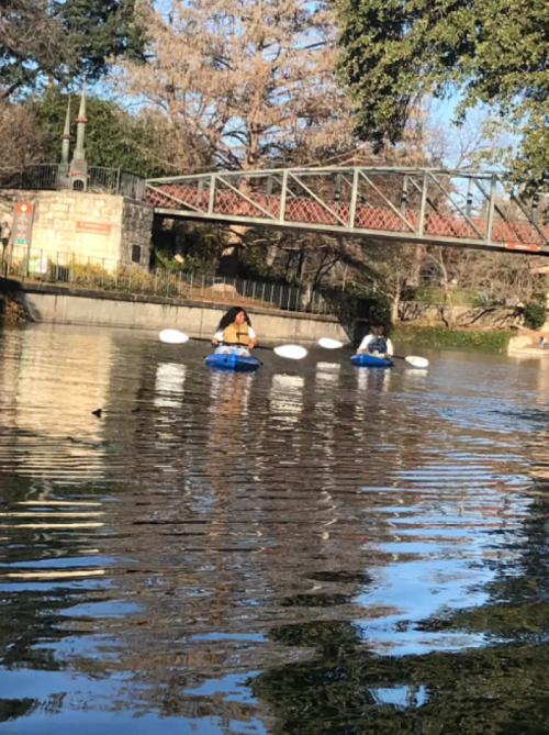 Two people in blue kayaks paddle on a calm river, with a bridge and trees in the background.