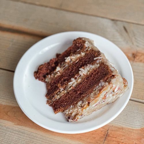 A slice of chocolate cake with coconut frosting on a white plate, resting on a wooden table.