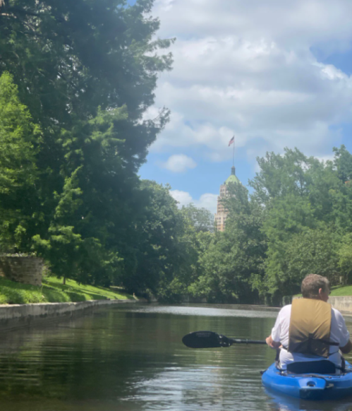 A person kayaking on a calm waterway, with trees and a building in the background under a partly cloudy sky.