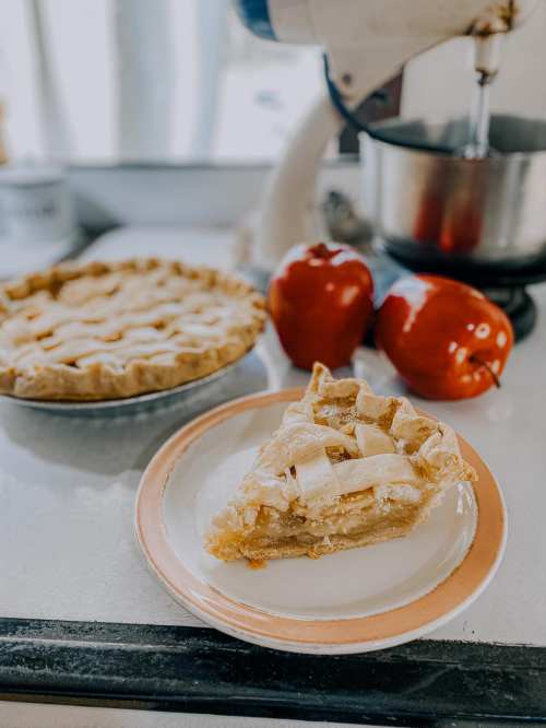 A slice of apple pie on a plate, with a whole pie and red apples in the background. A mixer is also visible.