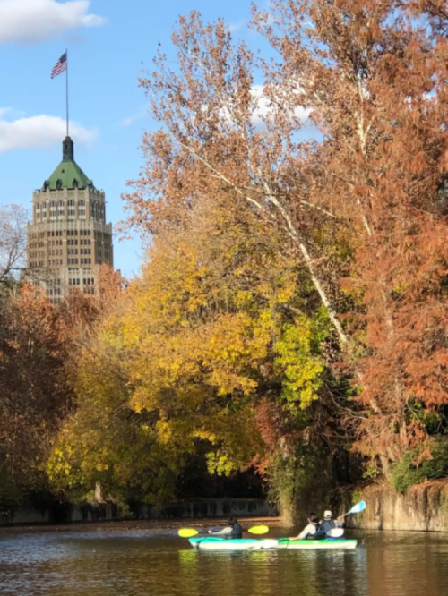 Two kayakers paddle on a calm river surrounded by autumn trees, with a tall building and American flag in the background.