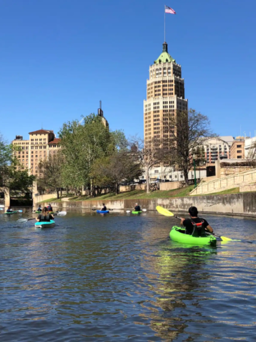 People kayaking on a river with city buildings and a clear blue sky in the background.