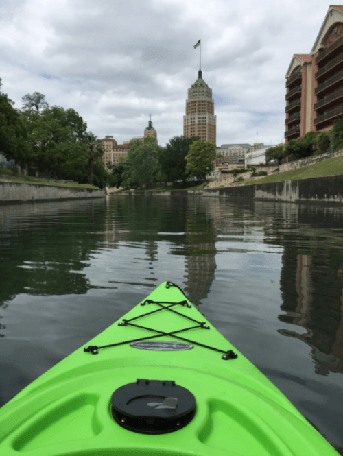 A green kayak on calm water, with city buildings and trees reflected in the surface under a cloudy sky.