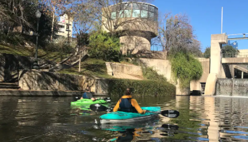 Two kayakers paddle on a calm waterway, with a waterfall and greenery in the background under a clear blue sky.