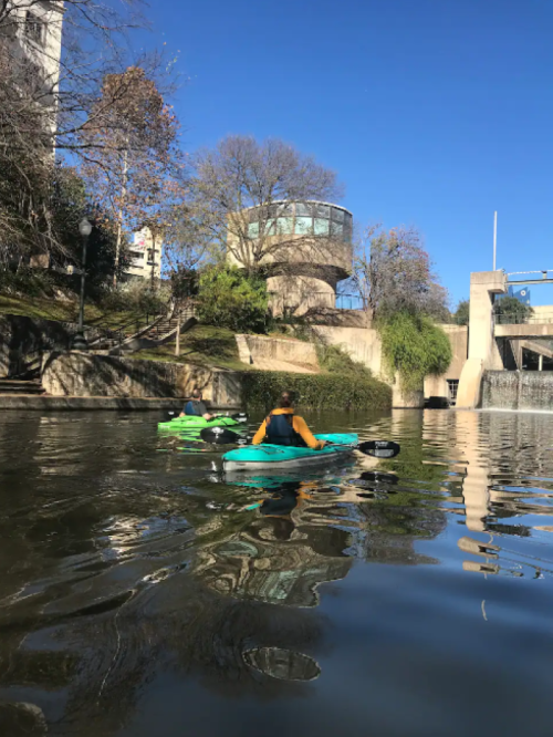Two kayakers paddle on a calm river, with trees and a modern building in the background under a clear blue sky.