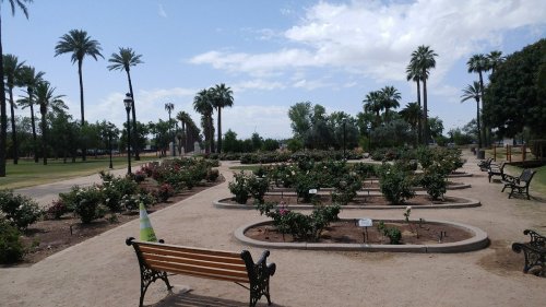 A park scene featuring flower beds, benches, and palm trees under a partly cloudy sky.