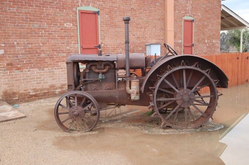 Rusty vintage tractor parked on gravel near a brick building with red shutters.