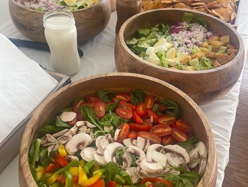 A table with wooden bowls of fresh salads, including greens, tomatoes, mushrooms, and croutons, with dressing nearby.