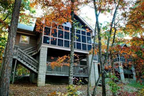 A wooden house surrounded by colorful autumn trees, featuring large screened windows and a staircase.