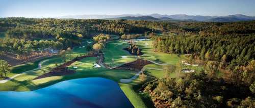 Aerial view of a lush golf course surrounded by trees and water, with mountains in the background under a clear sky.