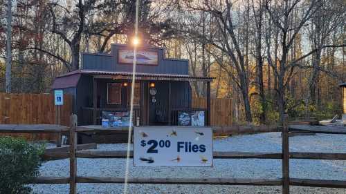 A small rustic shop with a sign advertising "$2 Flies" in a wooded area during sunset.