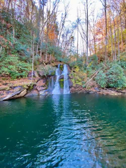 A serene waterfall cascades into a tranquil, greenish-blue pool, surrounded by lush trees and autumn foliage.