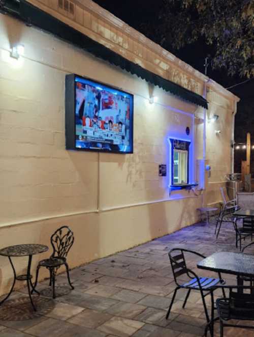 A cozy outdoor seating area with tables and a TV mounted on a wall, illuminated by soft blue lights at night.