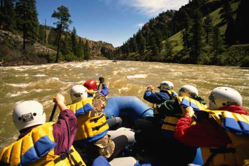 A group of people in helmets and life jackets paddling a raft through turbulent river waters surrounded by trees.