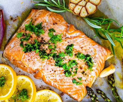 Baked salmon fillet topped with herbs, surrounded by lemon slices, garlic, and asparagus on parchment paper.