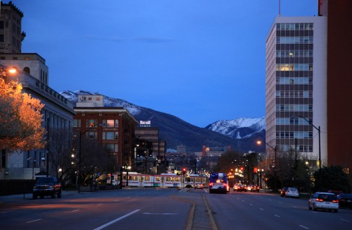 A city street at dusk, featuring buildings, a tram, and mountains in the background under a blue sky.