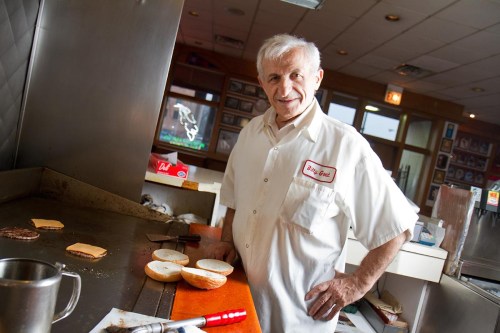 A smiling chef stands at a grill with burger patties and cheese, wearing a white shirt with a name tag.