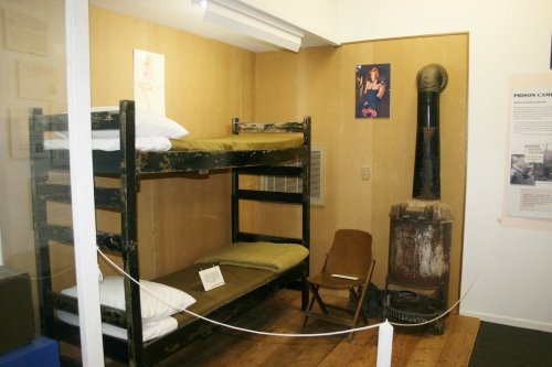 A simple museum display featuring a bunk bed, a chair, and a stove in a room with wooden flooring and wall art.
