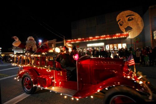 A festive parade featuring a vintage fire truck adorned with lights, Santa, and a large illuminated face in the background.
