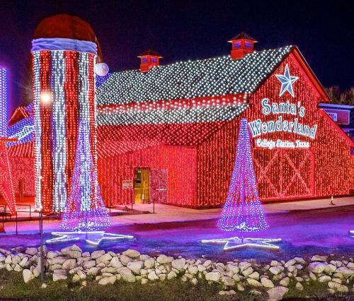 A brightly lit barn decorated with colorful Christmas lights, featuring festive trees and a large Santa hat on the roof.