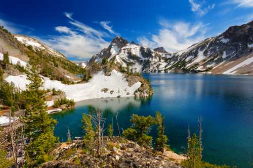 A serene mountain lake surrounded by snow-capped peaks and lush greenery under a bright blue sky.