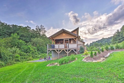 A modern cabin with a deck, surrounded by lush greenery and hills under a cloudy sky.