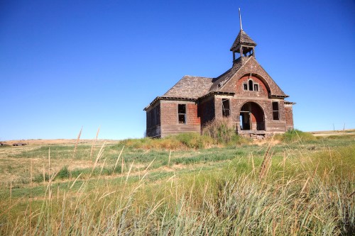 An old, abandoned schoolhouse with a bell tower, surrounded by tall grass under a clear blue sky.
