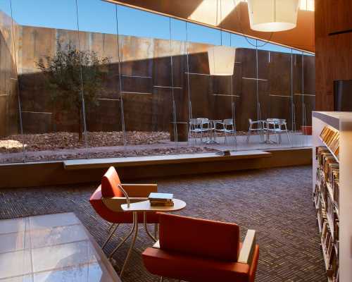 A modern library interior with red chairs, a small table, and large glass windows overlooking an outdoor space.