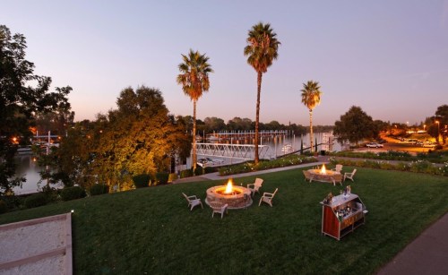 A serene riverside scene at dusk, featuring palm trees, fire pits, and a dock with boats in the background.