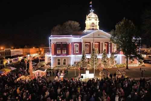 A festive town square at night, featuring a beautifully lit courthouse and a large crowd enjoying holiday decorations.