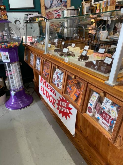 A wooden display counter filled with various fudge types, candy, and a gumball machine in a colorful shop.