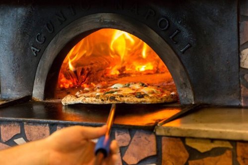A hand using a peel to remove a freshly baked pizza from a wood-fired oven with flames in the background.