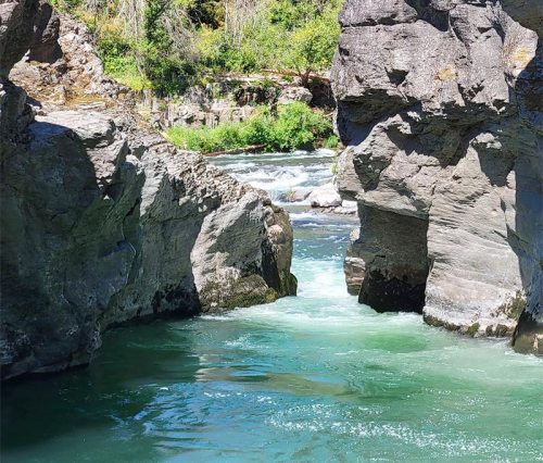 A serene river flows through a rocky landscape, with lush greenery in the background and clear blue water.