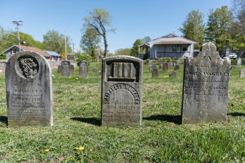 Three gravestones in a cemetery, surrounded by grass and trees, with a house visible in the background.