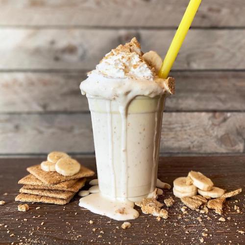 A creamy milkshake topped with whipped cream and graham cracker crumbs, served with a yellow straw and banana slices.