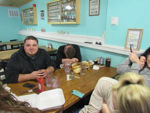 A group of friends at a restaurant table, with one person smiling and others laughing or resting their heads.
