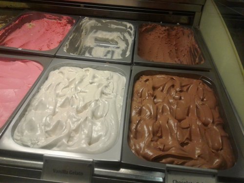 A display of gelato flavors, including pink, white, and brown varieties, in metal containers.