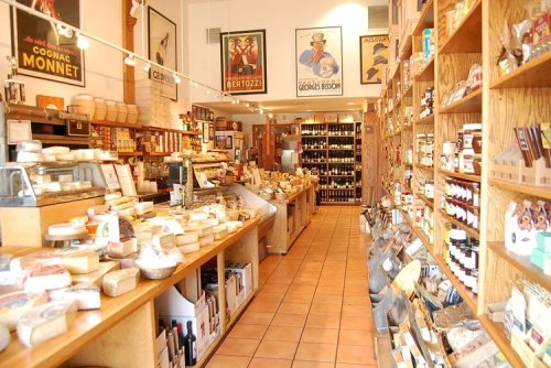 A cozy gourmet shop filled with shelves of cheese, jars, and various food products, featuring vintage posters on the walls.