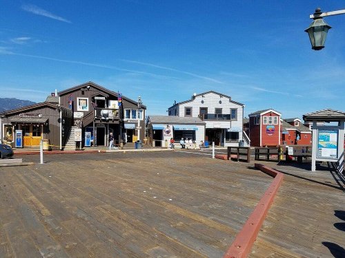 A sunny waterfront scene featuring colorful buildings and a wooden boardwalk. Clear blue sky above.