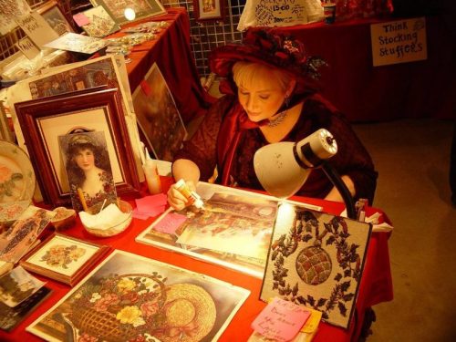 A woman in a red hat works on art at a table filled with framed pictures and crafts, illuminated by a lamp.