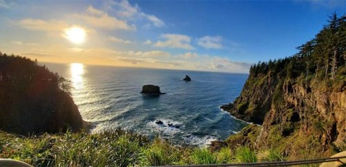 A scenic coastal view at sunset, featuring rocky cliffs, calm waters, and a bright sun reflecting on the ocean.