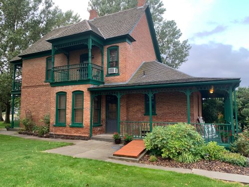 A two-story brick house with green trim, a porch, and a well-maintained lawn surrounded by trees.