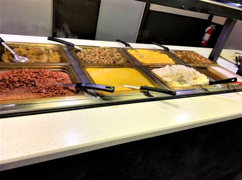 A buffet with various dishes, including beans, rice, and sauces, displayed in metal trays on a white countertop.