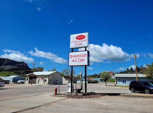 Sign for Uncle Louie's Diner with "Breakfast All Day" under a blue sky and scattered clouds. Nearby buildings visible.